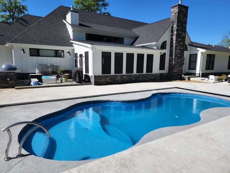 Maxima Pools long pool, rounded edges and curvy shaped, with a rail on the far left of the pool, surrounded by a concrete patio, bright blue sky, in front of a long white house with a lot of vertical windows and dark brick chimney.