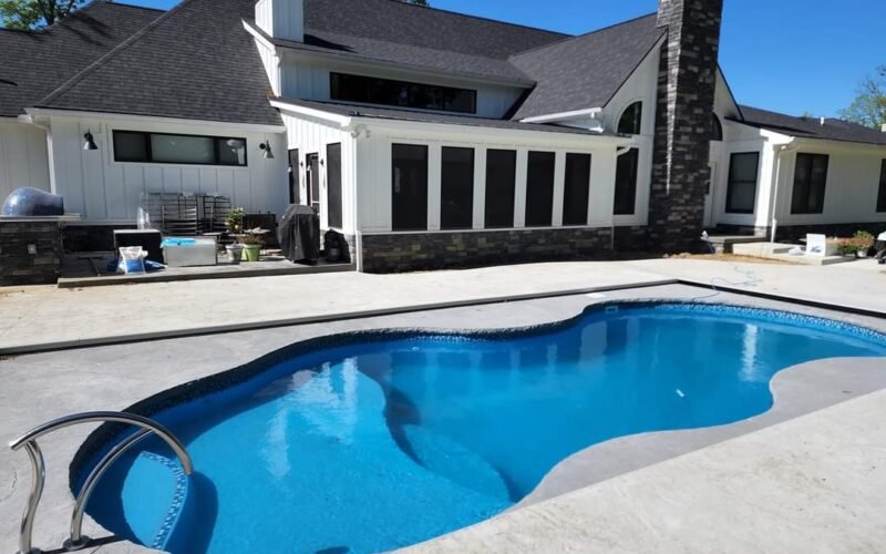 long pool, rounded edges and curvy shaped, with a rail on the far left of the pool, surrounded by a concrete patio, bright blue sky, in front of a long white house with a lot of vertical windows and dark brick chimney.