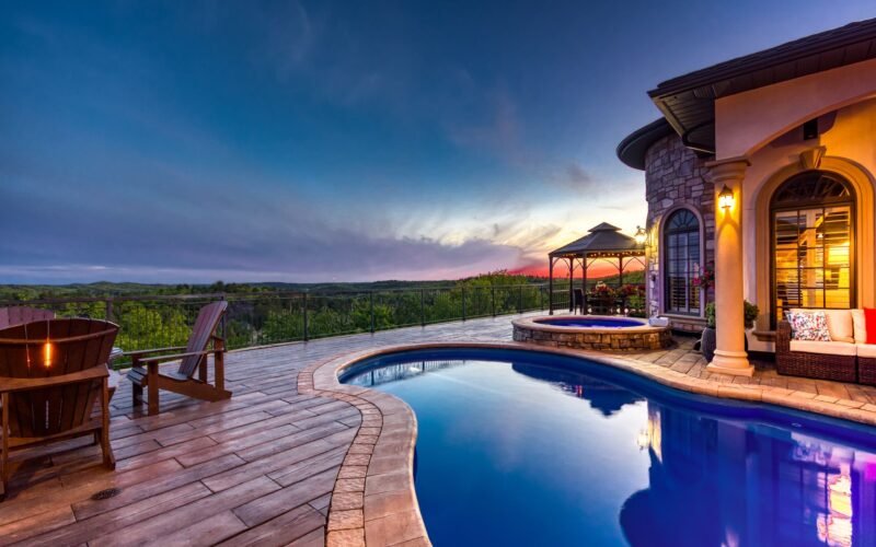 At dusk, dark blue sky with large thin clouds in the back, curvy pool righ in front of you dark blue water and a hot tub next to the far end of the pool on the right, in front of the house. light tan concrete around the pool, a concrete deck made to look like wood planks around it. light on hous pillar is on, three dark wood Adarondack chairs in the left corner of the deck, black iron fence at the back, looking slightly down on a forest of green trees.