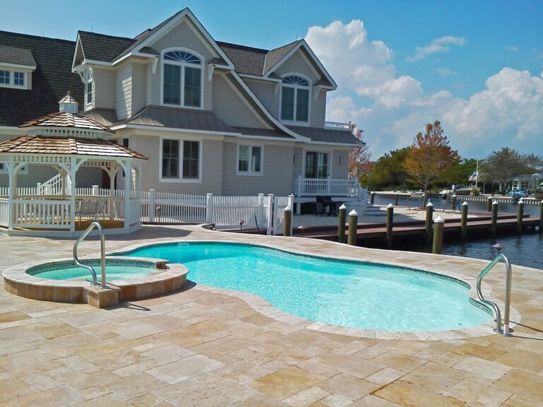 Maxima Pools Curvy pool with adjacent hot tub, tan concrete patio, white gazebo with a tan double roof to the far end of the pool, white picket fence behind it, beige two story house behind it, water way to the right of the pool and house. Blue sky with white fluffy clouds.