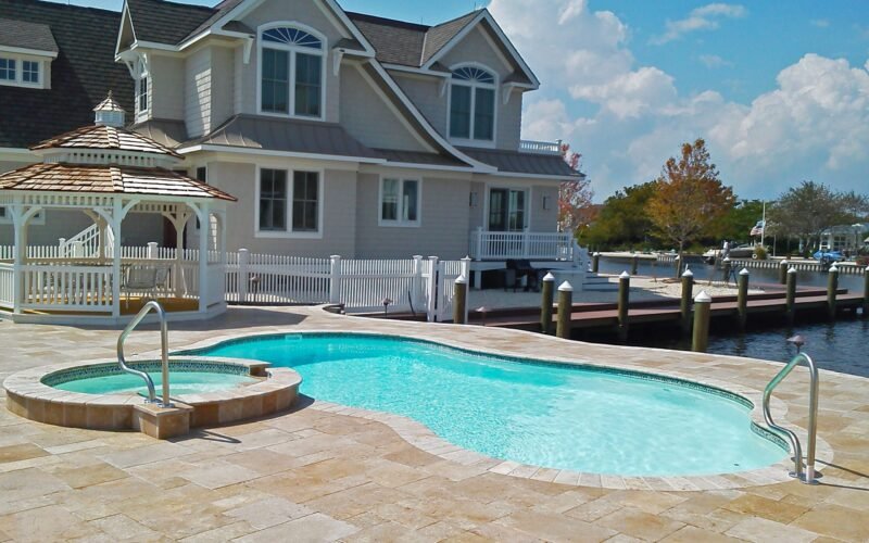 Curvy pool with adjacent hot tub, tan concrete patio, white gazebo with a tan double roof to the far end of the pool, white picket fence behind it, beige two story house behind it, water way to the right of the pool and house. Blue sky with white fluffy clouds.