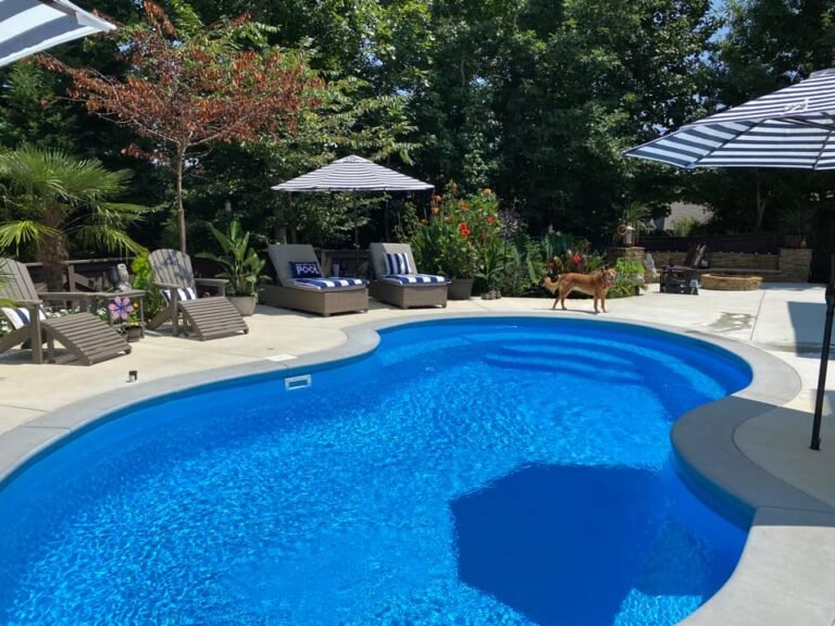 Maxima Pools Large, kidney-shaped pool, very blue water, a lot of green trees and foulage around the back of the pool, white concrete patio surrounding pool, three blue and white umbrellas around the pool with multiple lounge chairs and two Adirondack chairs on the left side, older tan, large dog next to pool with brick firepit in the back of the patio