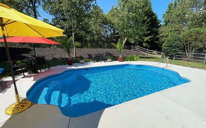 bright blue rounded pool in the middle of a large concrete patio, two large umbrellas to the left, one red and one yellow. Wooden fence on far right side, green trees around back and right hand side, large plants on the back side of the pool in front of small hill