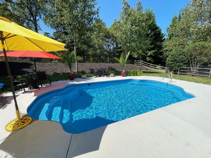 Maxima Pools bright blue rounded pool in the middle of a large concrete patio, two large umbrellas to the left, one red and one yellow. Wooden fence on far right side, green trees around back and right hand side, large plants on the back side of the pool in front of small hill