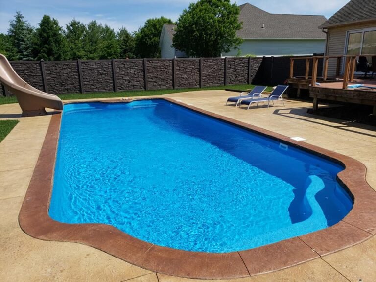 Maxima Pools long rectangular pool with rounded edges, blue water, sunny blue sky, slide on the far end left side of the pool, tan concrete patio, two blue lounge chairs on the right side of the pool, house with deck on far right. faux wood fence, green trees and white house in the far back