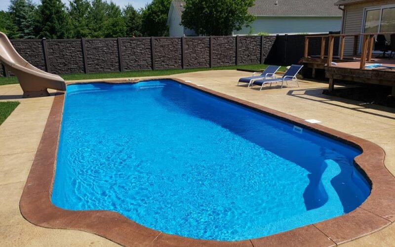 long rectangular pool with rounded edges, blue water, sunny blue sky, slide on the far end left side of the pool, tan concrete patio, two blue lounge chairs on the right side of the pool, house with deck on far right. faux wood fence, green trees and white house in the far back