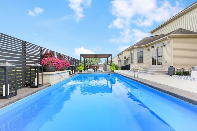 long angle of the rectangular pool, small waterfall on the left of the pool, from the pool water, tan house on the right. Pergola with seating area at the back of the screen, pink flowers in large round pots.