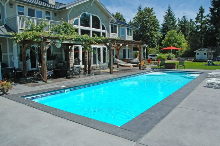 long rectangular San Juan Pool, crystal blue water, concrete patio with two pergolas in front of house with vines climbing. Sunny, blue sky with pine trees in the background.