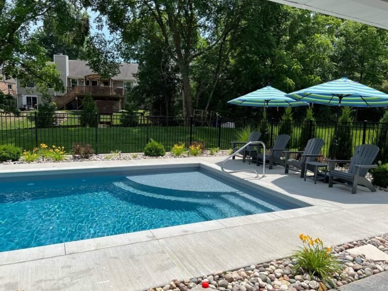 Maxima Pools rectangular pool with steps and railing on the left side, blue water, green yard with trees, grey Adirondack plastic chairs with two green and blue umbrellas. decorative rocks in the foreground, small plants and bushes around pool in front of black iron fence. neighbor's house in the background