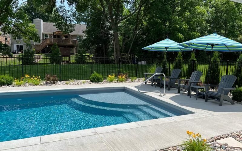 rectangular pool with steps and railing on the left side, blue water, green yard with trees, grey Adirondack plastic chairs with two green and blue umbrellas. decorative rocks in the foreground, small plants and bushes around pool in front of black iron fence. neighbor's house in the background