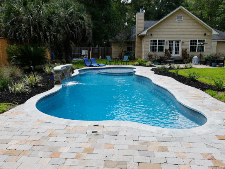Maxima Pools Long, curvy pool, blue water, small water fall on the left hand side, middle of the pool surrounded by a pool patio made of light gray and brown pavers. landscaping around the pool and green grass in front of a yellow house