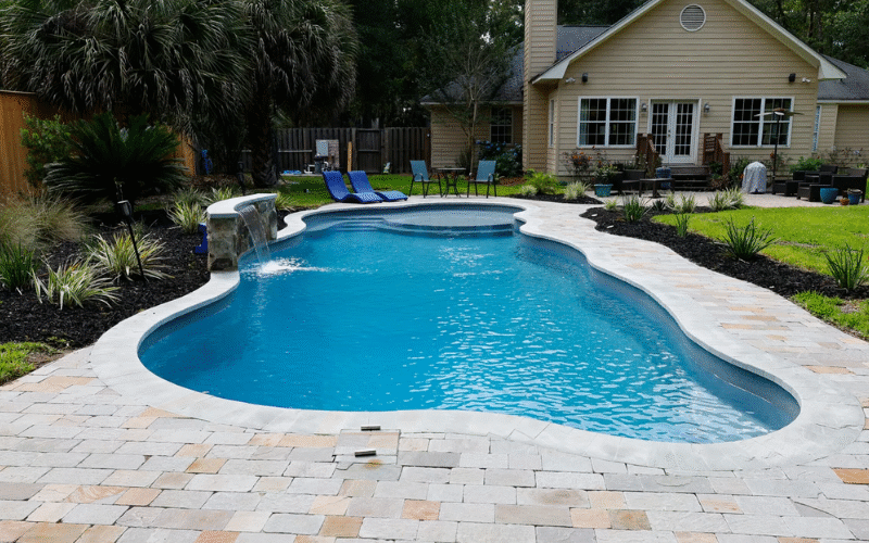Long, curvy pool, blue water, small water fall on the left hand side, middle of the pool surrounded by a pool patio made of light gray and brown pavers. landscaping around the pool and green grass in front of a yellow house