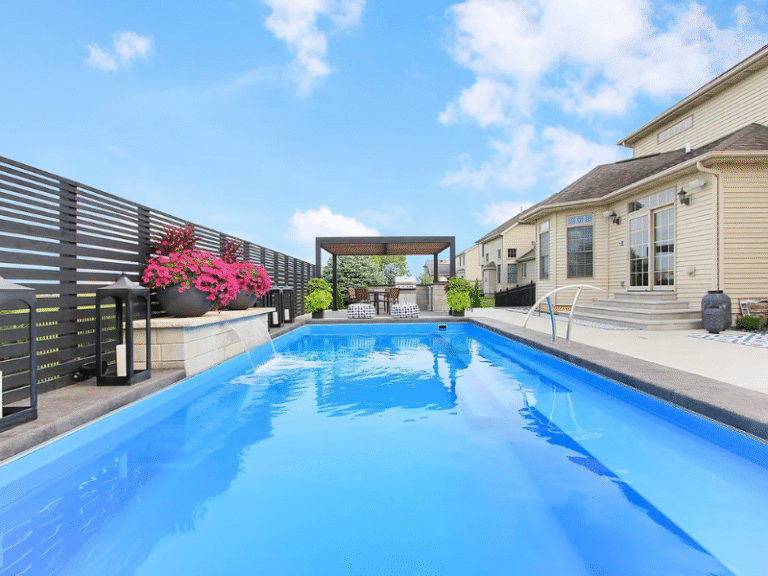 Maxima Pools long angle of the rectangular pool, small waterfall on the left of the pool, from the pool water, tan house on the right. Pergola with seating area at the back of the screen, pink flowers in large round pots.