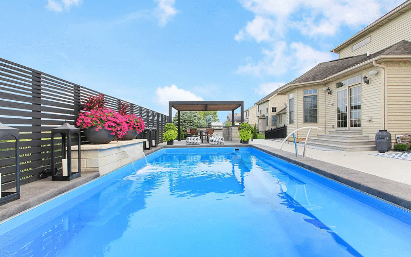 long angle of the rectangular pool, small waterfall on the left of the pool, from the pool water, tan house on the right. Pergola with seating area at the back of the screen, pink flowers in large round pots.
